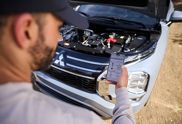 A man standing infront of a open Mitsubishi hood scheduling a service from his phone